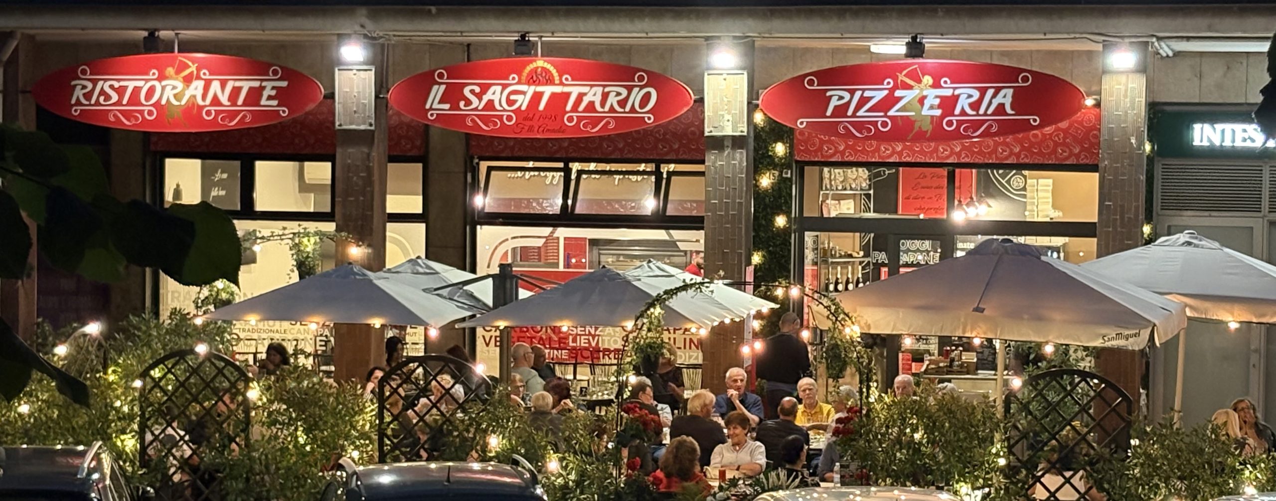 Esterno serale del ristorante pizzeria Il Sagittario con insegne rosse illuminate e tavoli all’aperto occupati dai clienti.